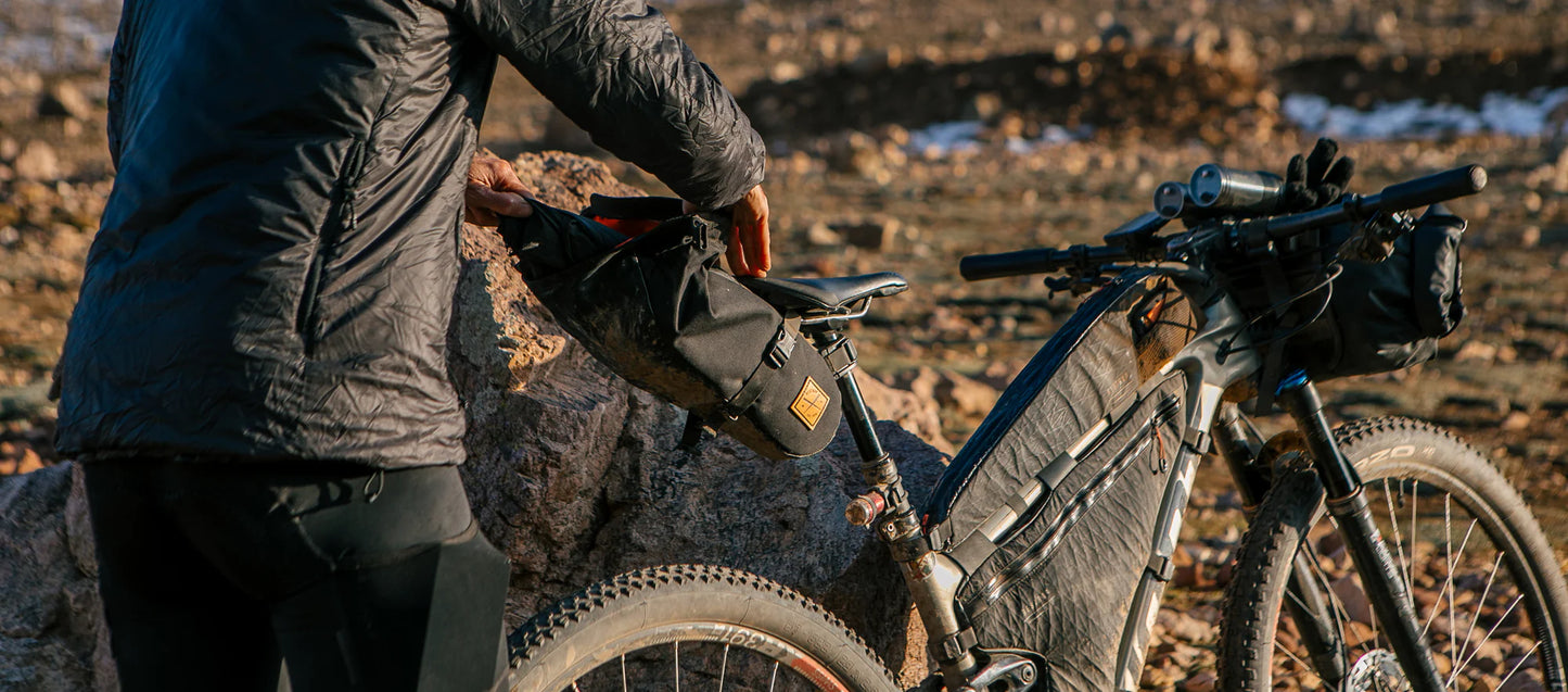 Een fietser in outdoor-kleding bevestigt een zwarte Restrap Saddle Pack aan het zadel van een bikepacking fiets in een ruig, rotsachtig landschap.