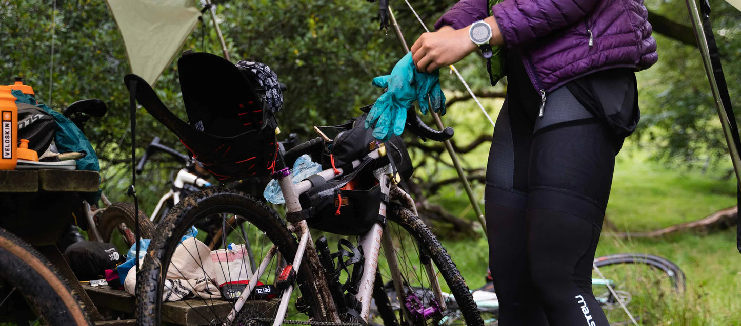 Een fietser is bezig met het inpakken van handschoenen in een Restrap frametas op een beboste kampeerplek.